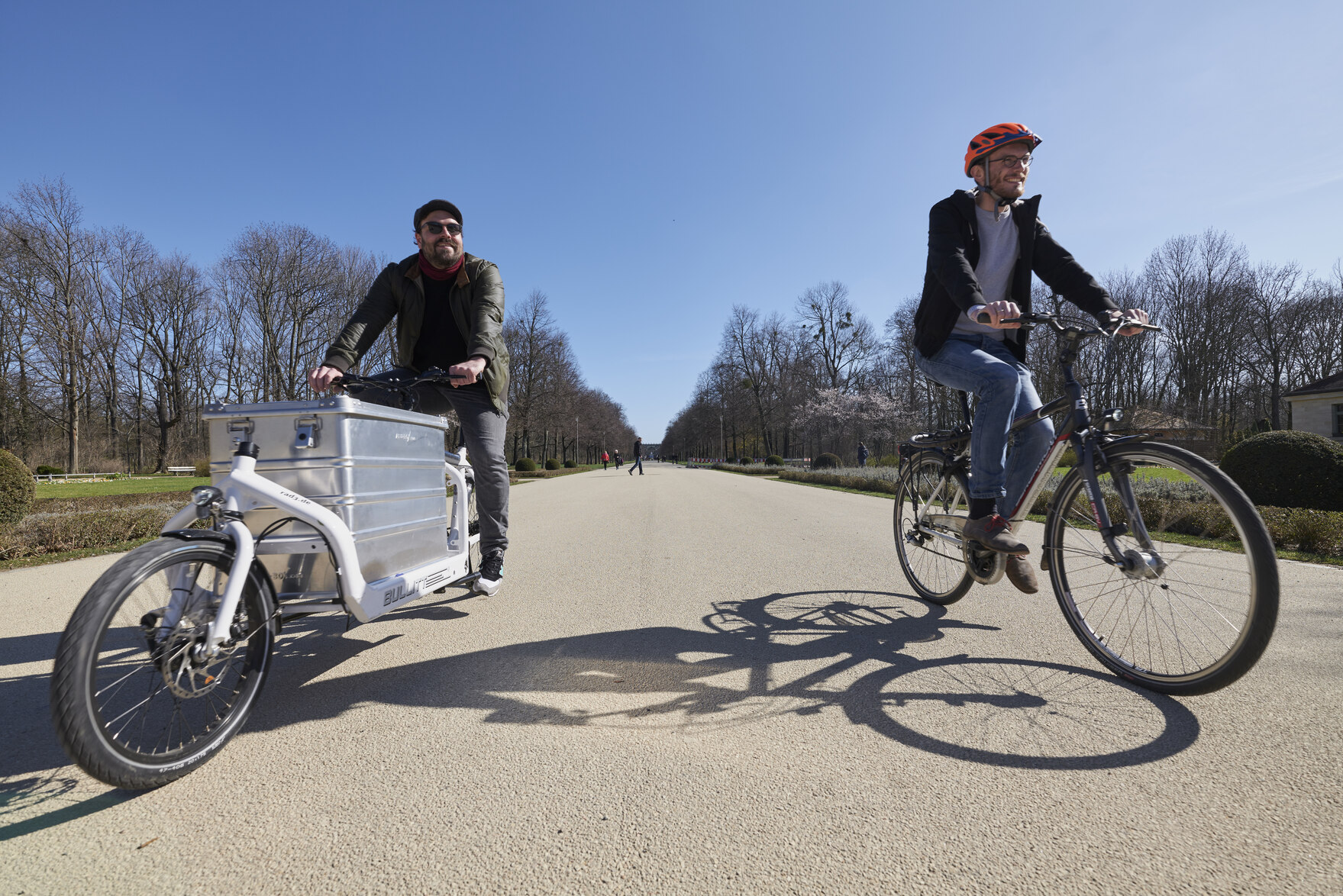 Ein Radfahrer auf dem Lastenrad und einer auf einem klassischen Fahrrad im Park.