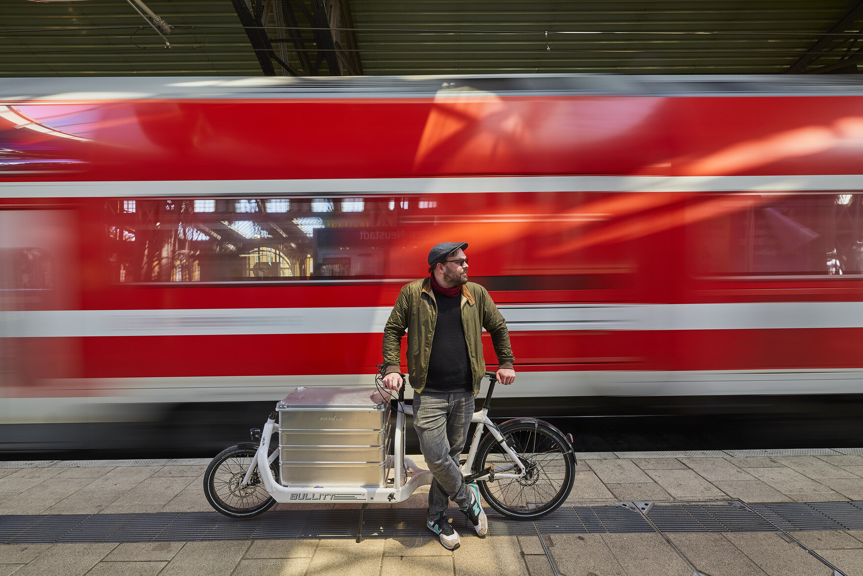 Ein Mann mit einem Lastenfahrrad steht vor einem fahrenden Zug.
